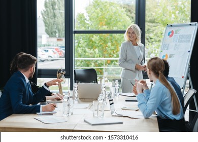 Smiling Businesswoman Standing Near Flipchart During Business Meeting With Young Colleagues