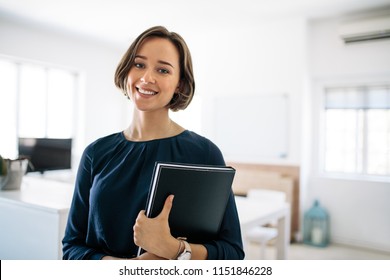 Smiling Businesswoman Standing In Office Holding A Diary. Portrait Of A Happy Woman Entrepreneur At Work.