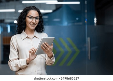 Smiling businesswoman standing in office using digital tablet. Professional looking confident while working with technology in modern workplace. Business and technology concept with office worker - Powered by Shutterstock