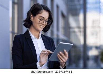 A smiling businesswoman is standing outside in an office center in a suit and using a tablet. Close-up photo. - Powered by Shutterstock