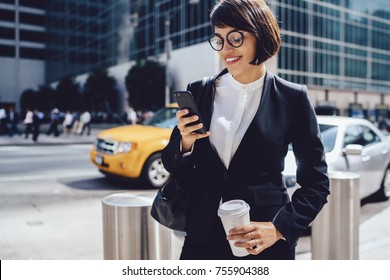Smiling businesswoman in stylish eyewear dialling number on mobile for calling taxi while standing on city street, prosperous female manager waiting for cab sending message and holding coffee to go