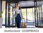 A smiling businesswoman is talking on her phone while pulling a suitcase through a hotel lobby. She appears confident and ready for her business trip, with natural light streaming through the windows.