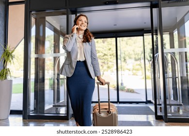 A smiling businesswoman is talking on her phone while pulling a suitcase through a hotel lobby. She appears confident and ready for her business trip, with natural light streaming through the windows.