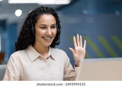 Smiling businesswoman using headset waving hand during online meeting in modern office environment. Female office worker engaged in video conference call, demonstrating positive communication - Powered by Shutterstock