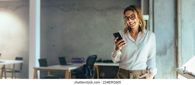 Smiling businesswoman using her phone in the office  Small business entrepreneur looking at her mobile phone   smiling while communicating and her office colleagues