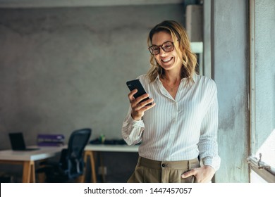 Smiling businesswoman using phone in office. Small business entrepreneur looking at her mobile phone and smiling. - Shutterstock ID 1447457057
