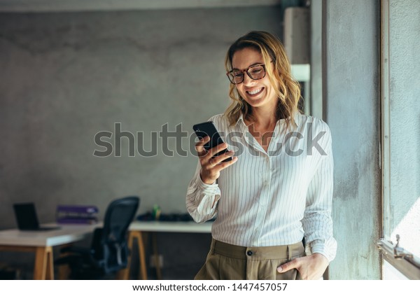Smiling\
businesswoman using phone in office. Small business entrepreneur\
looking at her mobile phone and\
smiling.