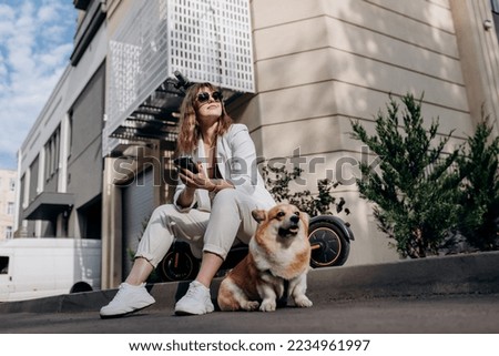 Smiling businesswoman in white suit sitting on electric scooter and using phone during walking with Welsh Corgi Pembroke dog in city