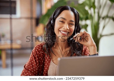 Smiling businesswoman working in a creative office. Smiling friendly woman working as call center agent for online support. Mixed race girl in video call with headphones looking at camera.