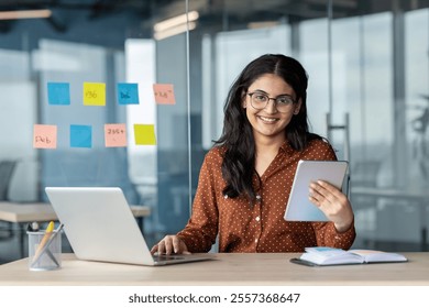 Smiling businesswoman working in office with a laptop and tablet, highlighting productivity and success. Professional setting portrays a positive and capable demeanor ready to achieve business goals. - Powered by Shutterstock