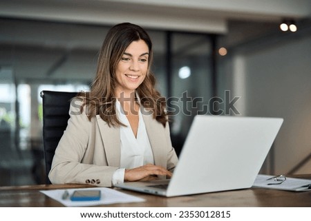 Photo of Smiling busy mature middle aged professional business woman manager executive wearing suit looking at laptop computer technology in office working on digital project sitting at desk.