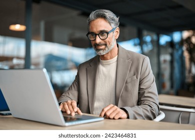 Smiling busy middle aged mature professional business man entrepreneur wearing suit and eyeglasses sitting at table outdoors working online using laptop computer hybrid working outside office. - Powered by Shutterstock