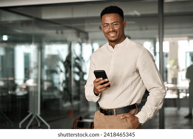 Smiling busy professional African American business man standing in office holding mobile cellphone. Young Black businessman entrepreneur using smartphone looking at camera with cell phone at work. - Powered by Shutterstock