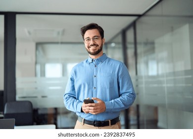 Smiling busy professional latin business man standing in office holding mobile cellphone. Young happy businessman employee using smartphone looking at camera using cell phone tech at work. Portrait.