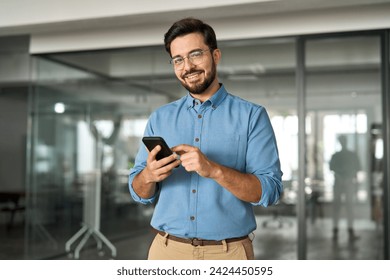 Smiling busy professional latin business man standing in office holding mobile cellphone. Young happy businessman employee using smartphone looking at camera using cell phone tech at work. Portrait.