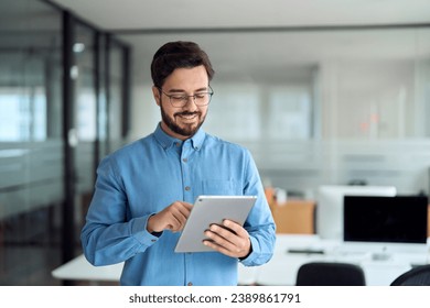 Smiling busy young latin business man manager using tablet computer, happy hispanic businessman executive looking at tab device analyzing finance trading market data working standing in office.