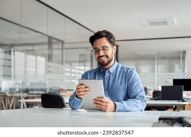 Sonriente joven y ocupado gerente de negocios latino usando tablet computer, feliz empresario hispano ejecutivo mirando el dispositivo de tabulación analizando los datos del mercado de comercio financiero trabajando en oficina.
