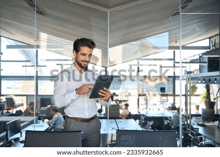 Smiling busy young Latin business man entrepreneur using tablet standing in office at work. Happy male professional executive manager using tab computer managing financial banking tech data.