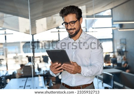 Smiling busy young Latin business man entrepreneur using tablet standing in office at work. Happy male professional executive manager using tab computer managing financial banking or marketing data.