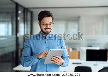 Smiling busy young latin business man manager using tablet computer, happy hispanic businessman executive looking at tab device analyzing finance trading market data working standing in office.