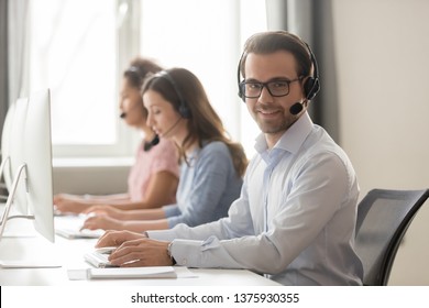 Smiling call center male worker pose looking at camera girls colleagues on background, sales agent wearing headset use pc answers incoming phone calls, provide professional support to customer concept