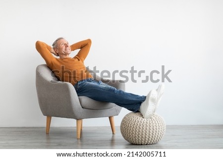 Smiling calm mature man relaxing sitting on armchair, resting feet on knitted pouf, free copy space. Happy adult spending weekend, leaning back holding hands behind head, isolated on white studio wall