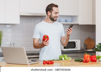 Smiling Calm Young Unshaven Housewife Man 20s In Casual Basic Blue T-shirt Using Mobile Phone, Looking At Recipe Hold Bell Pepper Preparing Vegetable Salad Cooking Food In Light Kitchen At Home Alone.