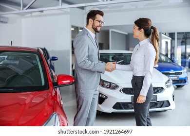 Smiling car seller shaking hands with brunette who wants to buy a car. Car salon interior. All around are many different modern cars.