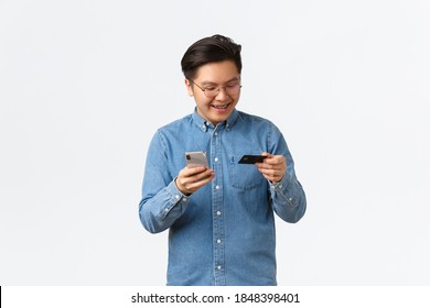 Smiling Carefree Asian Man In Glasses And Casual Clothes, Having Braces, Looking At Credit Card Pleased, Buying Online With Smartphone App, Shopping In Internet With Mobile Phone