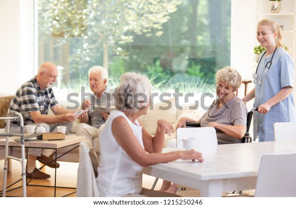 Smiling caregiver supporting happy elderly\
woman in the wheelchair in the nursing\
house