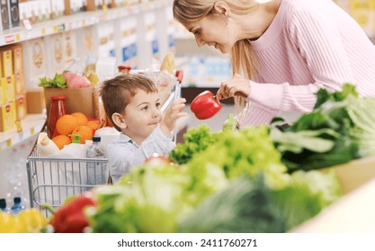 Smiling caring mother and cute kid doing grocery shopping together, they are picking vegetables in the produce section at the supermarket - Powered by Shutterstock