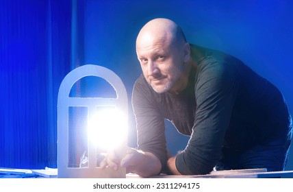 Smiling carpenter. Man with miniature window frame. Guy is showing off detail for cottage model. Happy carpenter near table with boards. Manufacture wooden furniture. Man carpenter looking at camera - Shutterstock ID 2311294175