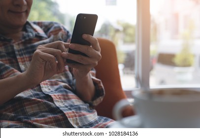 Smiling Casual Asian Man Or Hipster Leaning On Armchair Relaxed Messaging On Mobile Smart Phone With Cup Of Coffee On Table Waiting For Someone In Cafe Or Coffee Shop, Using Application On Phone