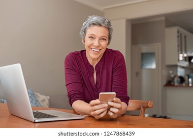 Smiling Casual Senior Woman Using Laptop While Messaging With Smartphone. Happy Mature Woman Working With A Cellphone And Laptop At Home And Looking At Camera. Business Woman Using Her Mobile Phone.