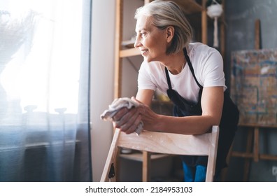 Smiling caucasian 50 years old female sculptor cleaning hands with rag after making clay. Small business and entrepreneurship. Home hobby, entertainment and leisure. Woman looking away. Art studio - Powered by Shutterstock