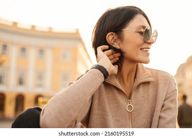 Smiling caucasian adult woman with dark hair walking on sunny street. Beauty wears casual jacket and sunglasses. People sincere emotions lifestyle concept.