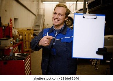 Smiling Caucasian Auto Mechanic, Car Engineer, Technician Pointing Blank Clipboard Sheet In Hand With Copy Space For Car Repair Ad Checklist While Standing Against Car Lifts In The Repair Shop Garage