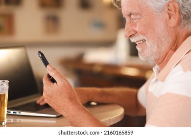 Smiling caucasian bearded senior man sitting at pub table with a glass of beer working on laptop using phone, addicted elderly  enjoying tech and social - Shutterstock ID 2160165859