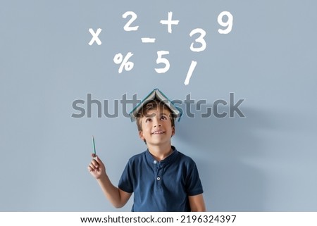 Smiling Caucasian boy with a book on his head pointing to various mathematical symbols with a pencil. Back to school, time to study subjects with numbers