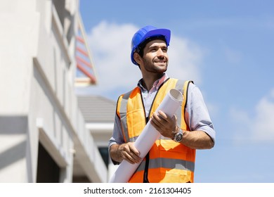 A smiling caucasian builder in a white hard hat and yellow fluorescent jacket holds clipboard, stands on the roof, inspects construction site and makes notes.