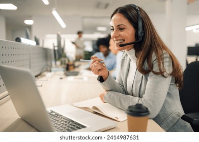 Smiling caucasian business woman with headset working on a laptop in a call center on the line. Happy female working in a busy customer service department and taking notes while talking with a client. - Powered by Shutterstock