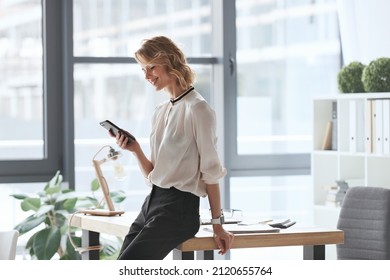 Smiling caucasian businesswoman watching on smartphone during work at office. Concept of modern successful woman. Idea of business and entrepreneur lifestyle. Young beautiful woman standing at table - Powered by Shutterstock