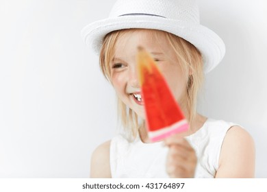 Smiling Caucasian Child Laughing And Hiding Behind Red Popsicle In Her White Beachwear Summer Clothes. Small Blond Girl Looking Sideways In Playful Manner Of Happy-go-lucky Kid.