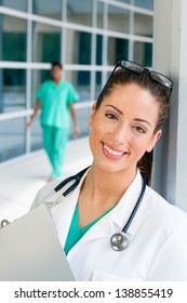 Smiling Caucasian doctor holding a clipboard in brightly lit exterior hospital environment in scrubs, white lab coat and holding glasses. Nurse in scrubs in the background.