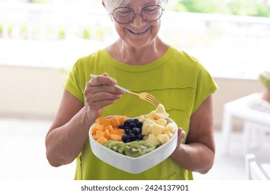 Smiling caucasian elderly woman holding a heart shape bowl with a fresh ready-to-eat summer fruit salad, kiwi, melon, apple, blueberries. Healthy eating concept - Powered by Shutterstock