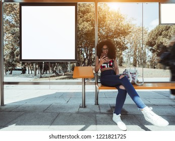 A smiling caucasian female with bulky curly hair and in eyeglasses is sitting inside of the outdoor bus stop and using her smartphone with an empty white mockup of information billboard next to her - Powered by Shutterstock