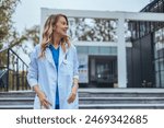 A smiling Caucasian female healthcare worker stands relaxed outside a clinic, her hands resting casually in her lab coat pockets.