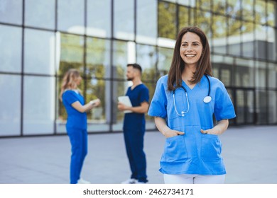 A smiling Caucasian female nurse in blue scrubs with a stethoscope stands confidently outside a modern hospital, with colleagues in the background. - Powered by Shutterstock