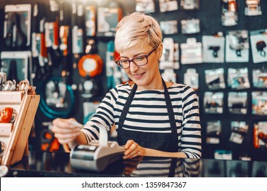 Smiling Caucasian female worker with short blonde hair and eyeglasses using cash register while standing in bicycle store.