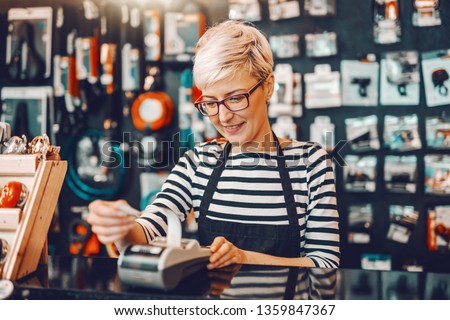 Smiling Caucasian female worker with short blonde hair and eyeglasses using cash register while standing in bicycle store.